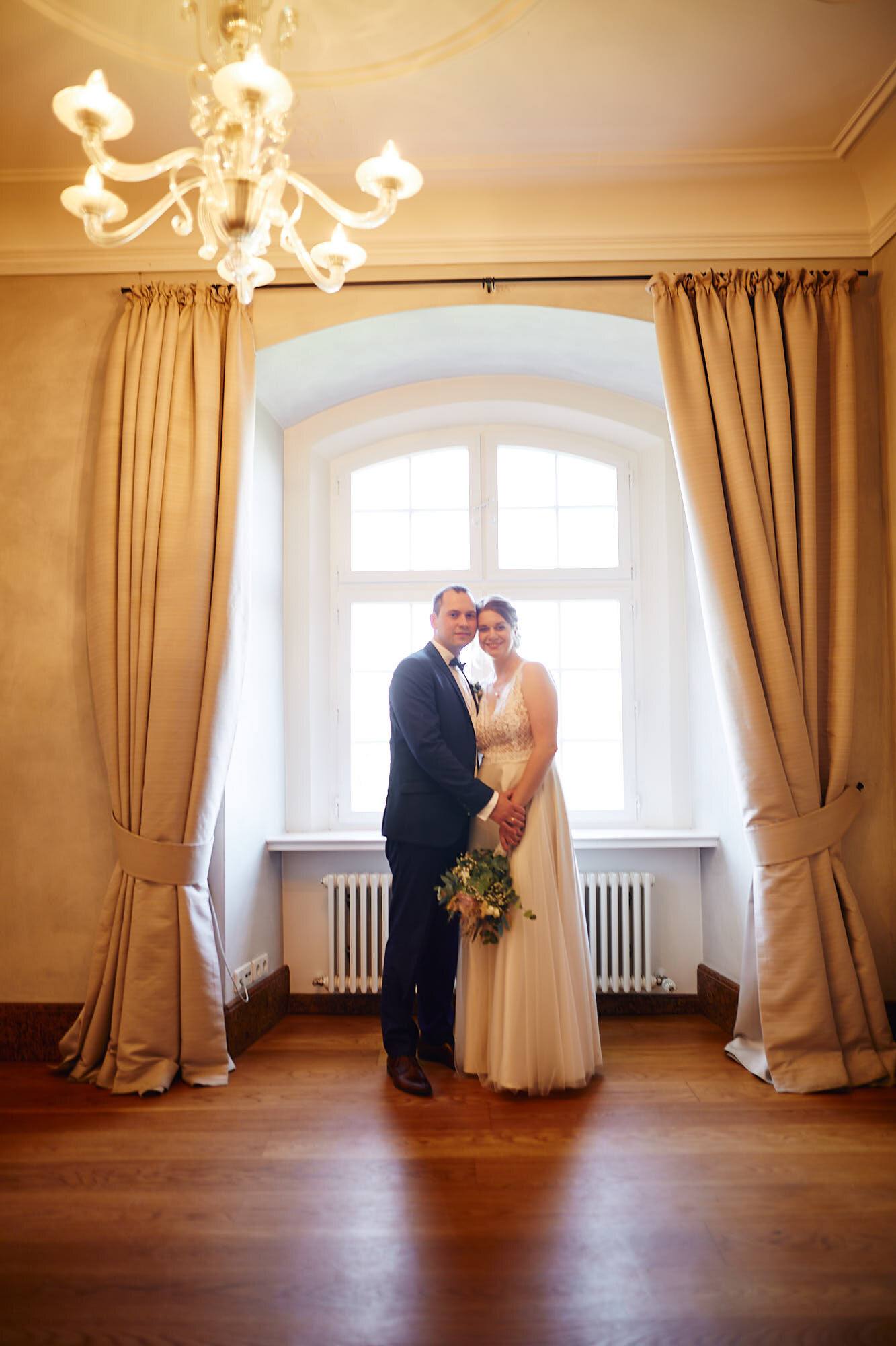 Brautpaar im Salon vor hohem Fenster mit goldenen Vorhängen und Kronleuchter