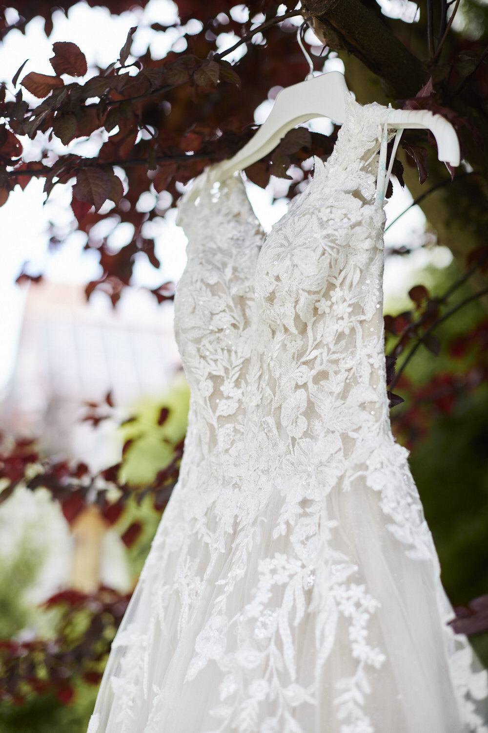 Brautkleid mit floraler Spitze hängt im Gegenlicht vor einem Baum mit rotem Laub