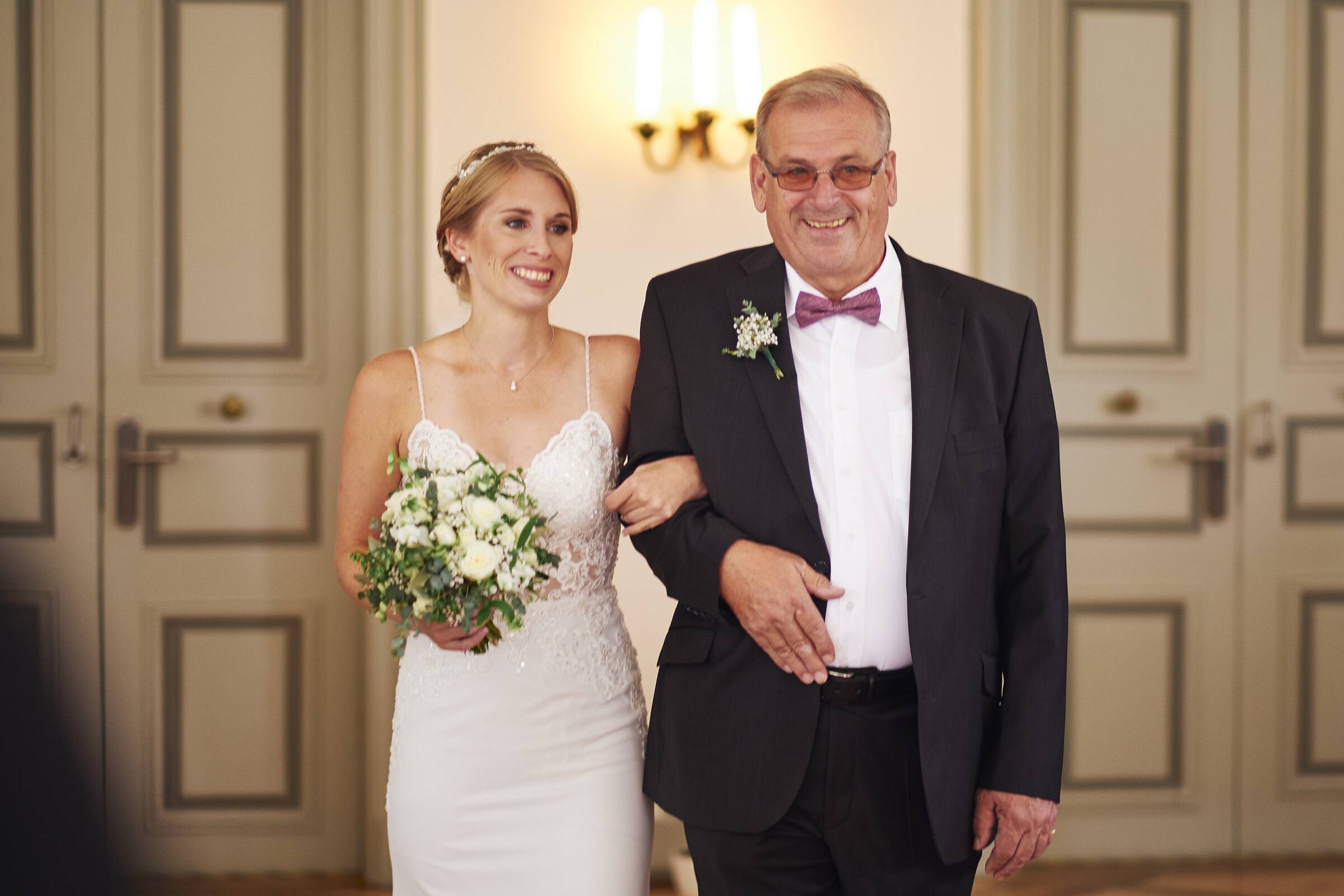 Braut und ihr Vater lächeln vor der Flügeltür kurz vor dem Einzug