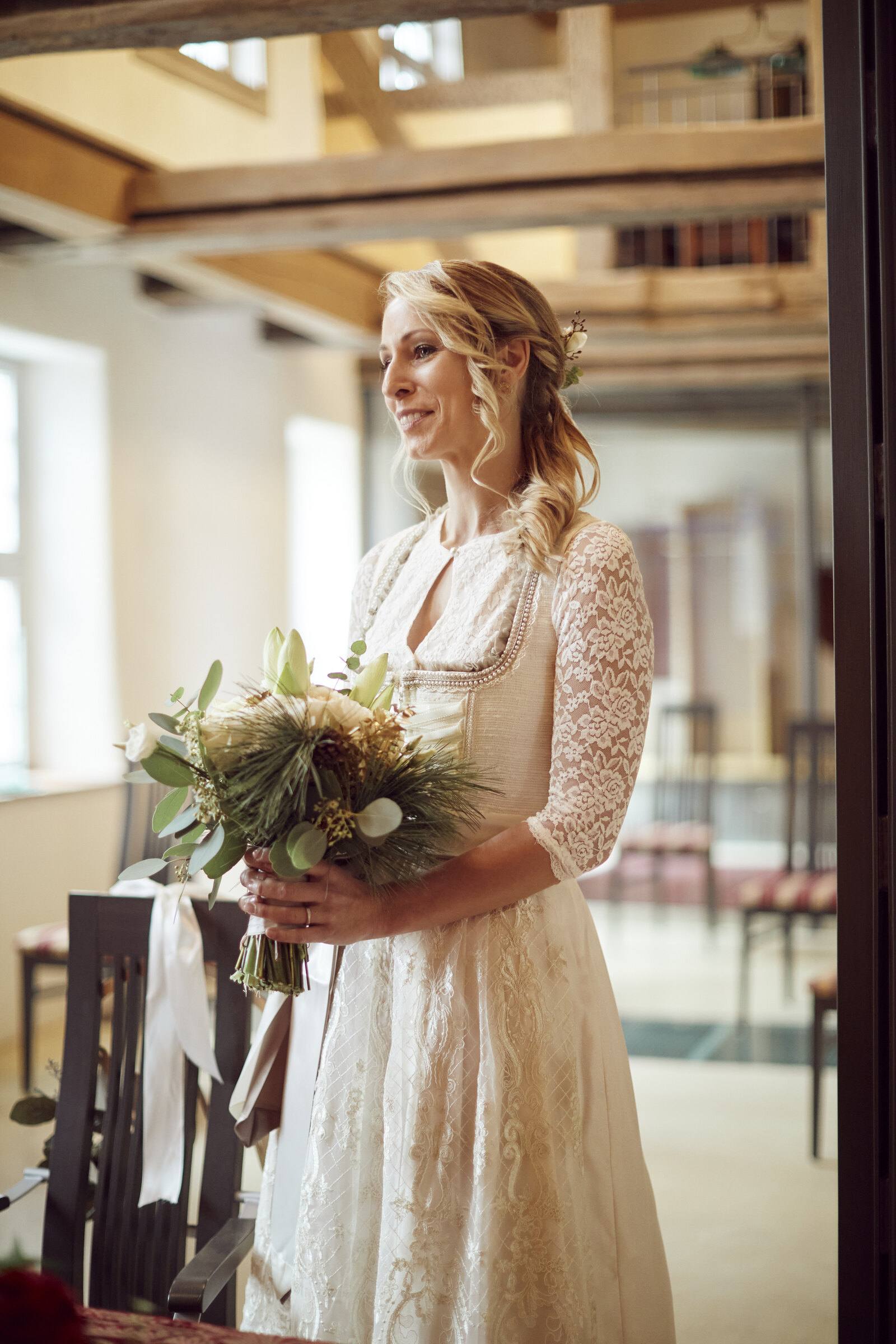 Braut im cremefarbenen Spitzendirndl mit Strauß im Trauzimmer mit Holzbalken