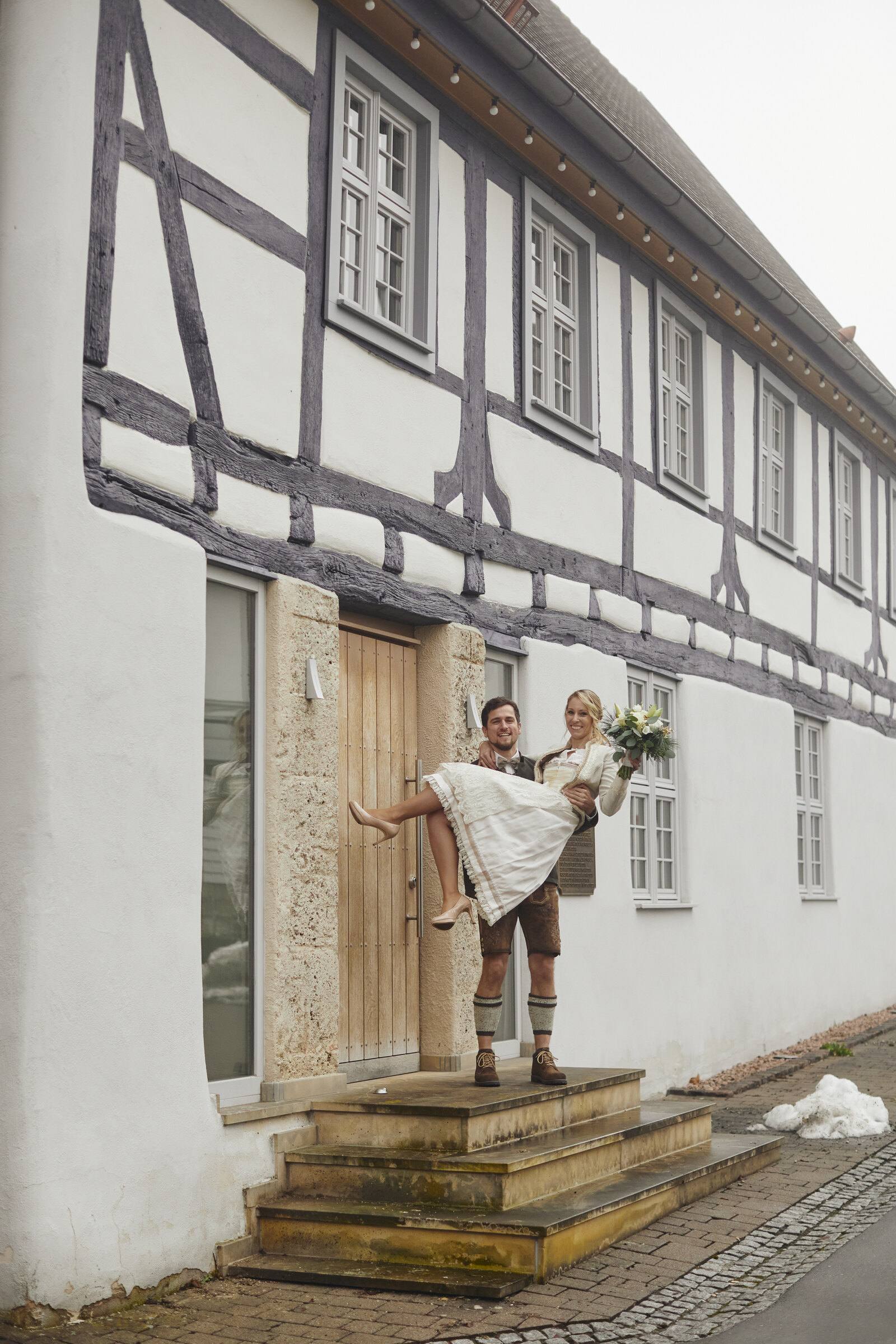 Bräutigam in Lederhose trägt die Braut vor einem Fachwerkhaus