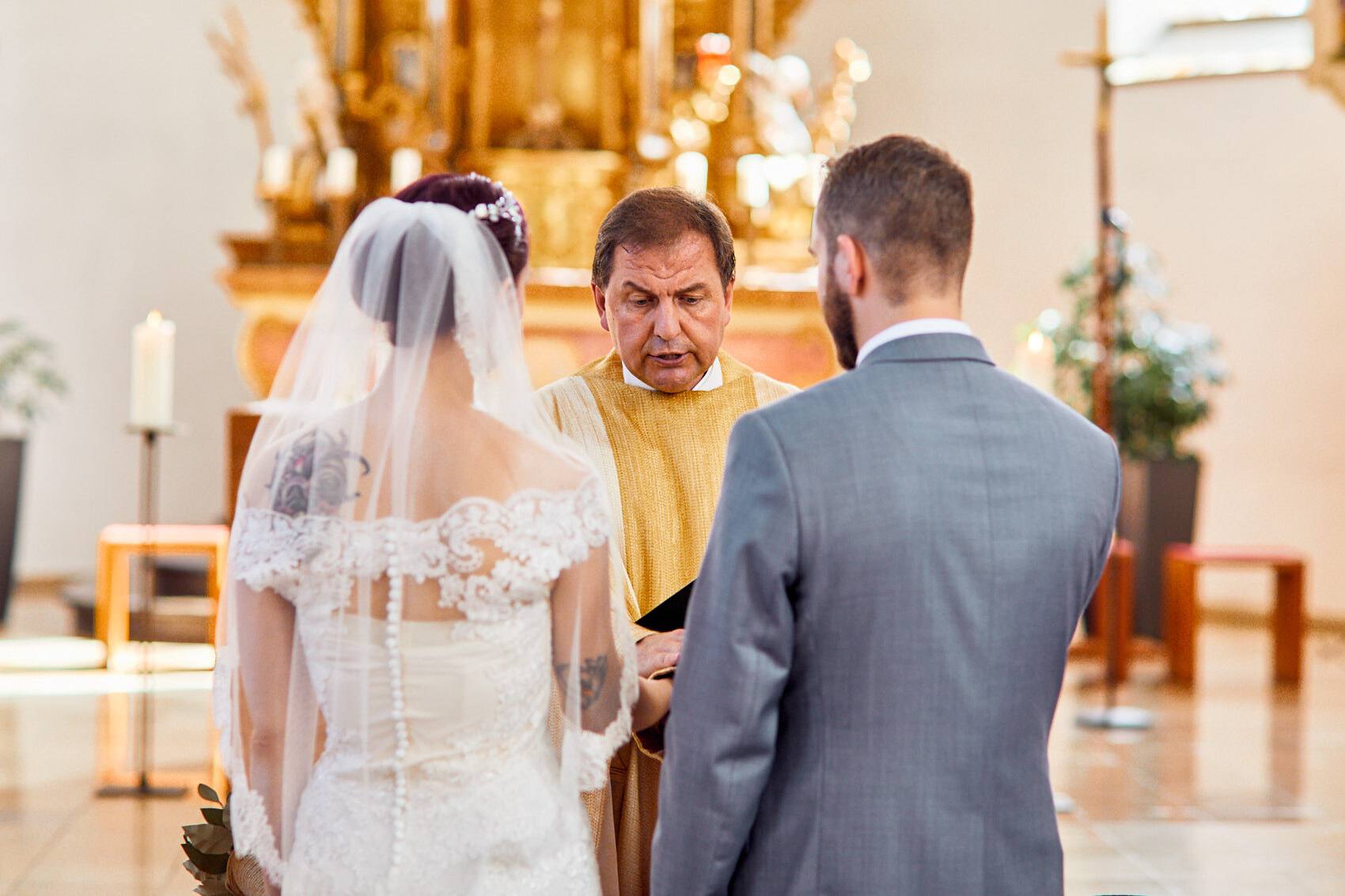 Braut mit Spitzenkleid und Schleier und Bräutigam vor dem goldenen Altar mit Pfarrer