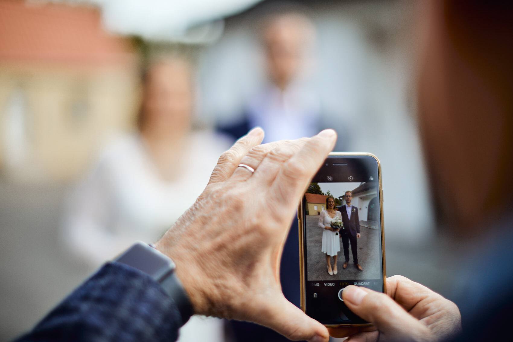 Gast fotografiert das Brautpaar mit dem Smartphone, das Paar unscharf im Hintergrund