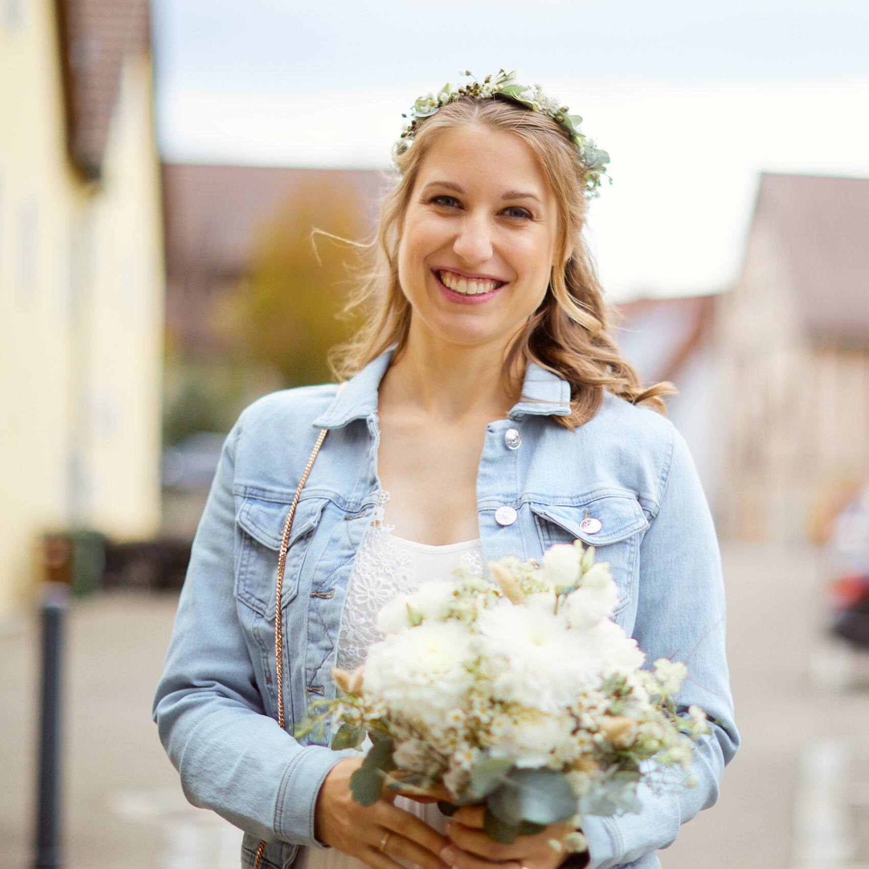 Lachende Braut mit Blumenkranz und Jeansjacke hält ihren Strauß