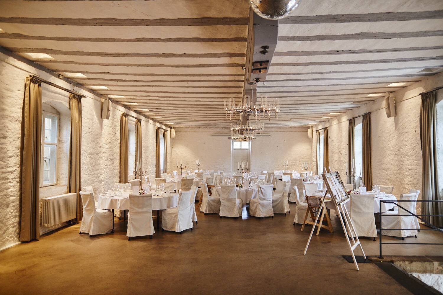 Festsaal mit Holzbalkendecke und weiß gedeckten Tischen.