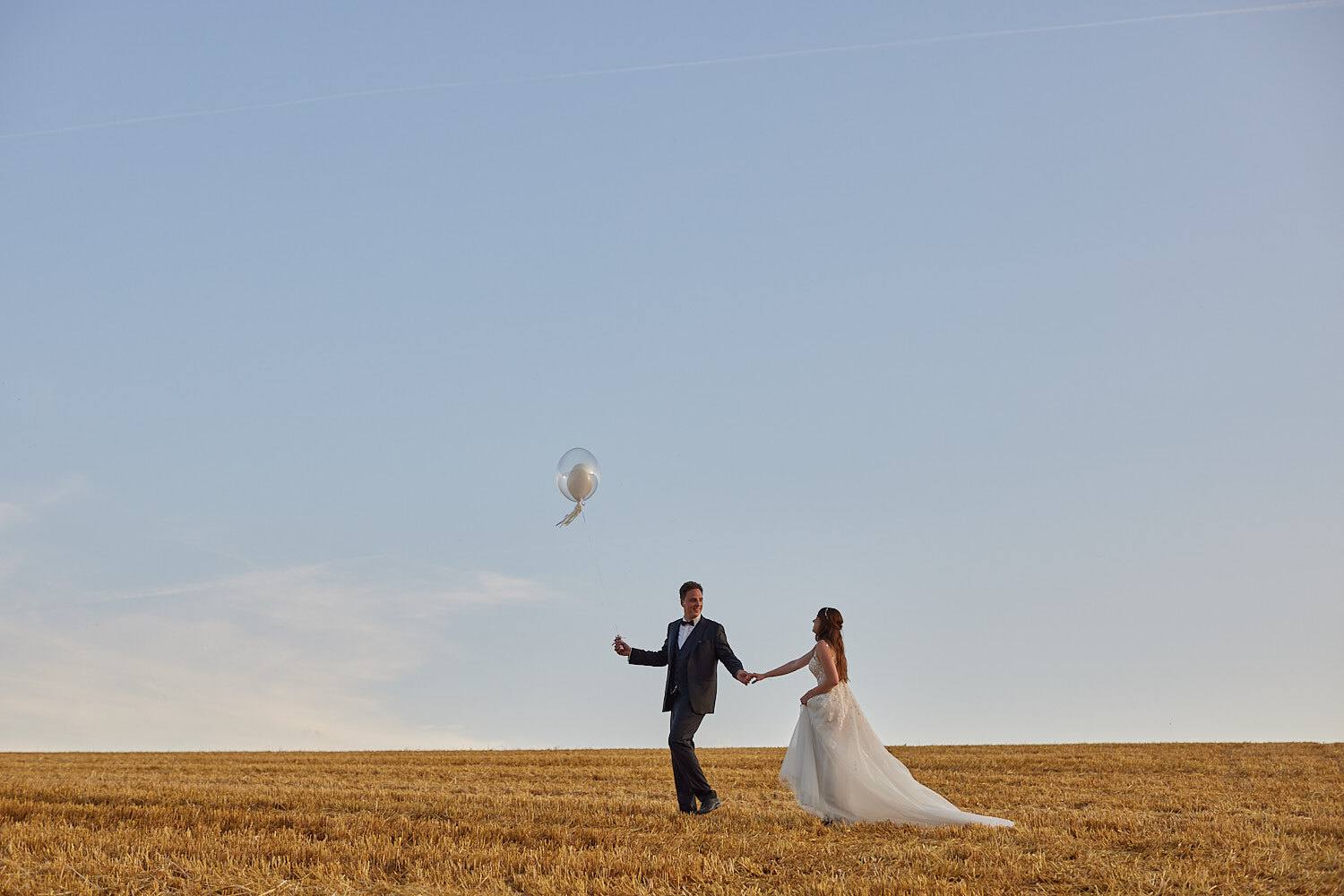 Brautpaar Hand in Hand im Feld, ein silberner Ballon schwebt in den Himmel